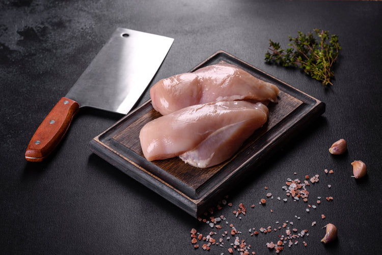 chicken breast on wood chopping block, next to cleaver, with black background